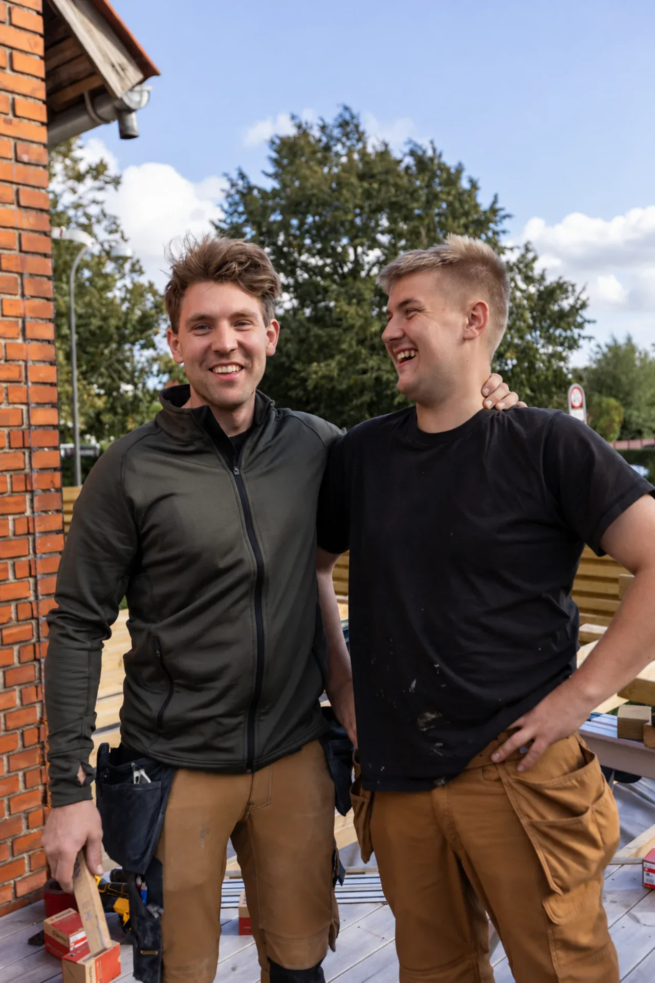 To mænd smiler og snakker udenfor, med træer og en bygning i baggrunden.